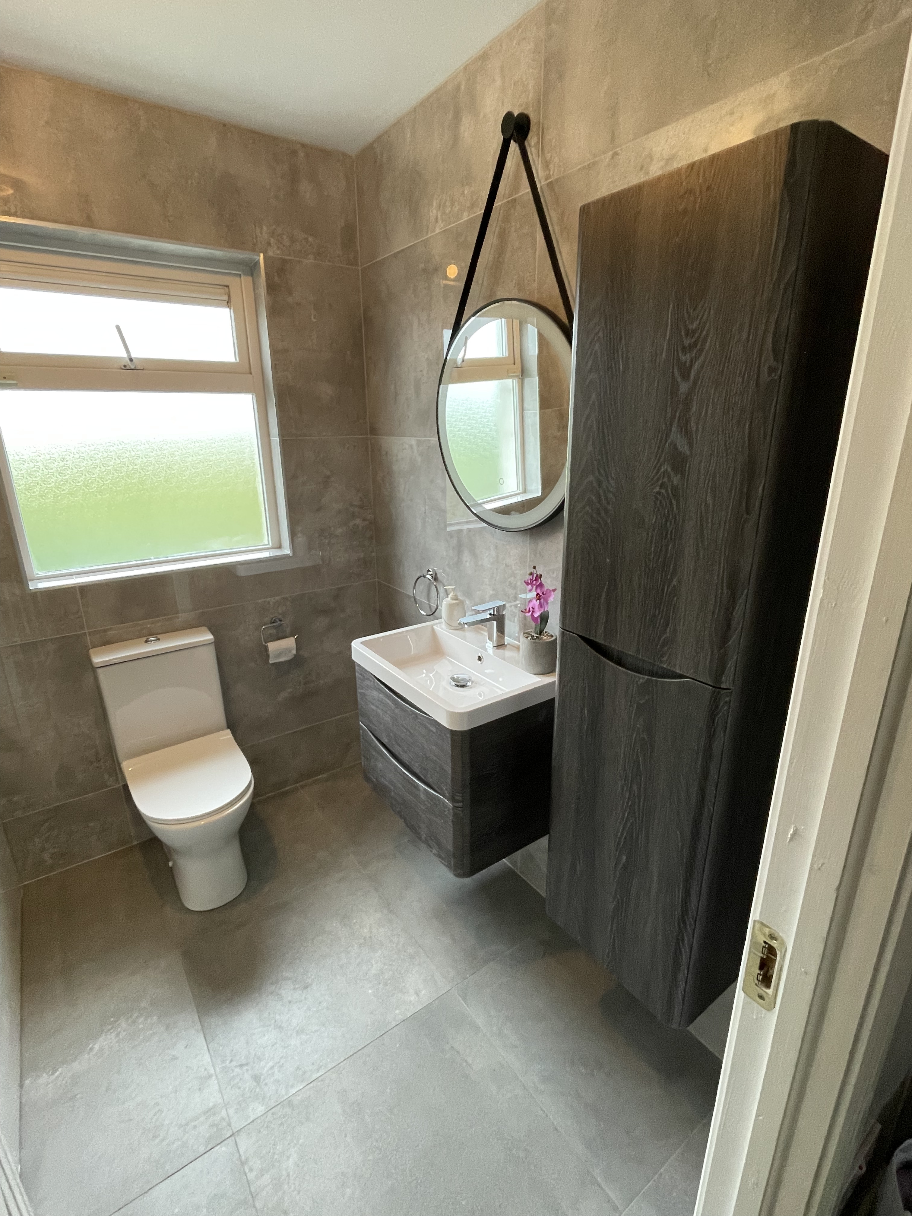 Custom built bathroom with brown furniture a white toilet. Hanging mirror above a brown floating sink and window in the background.