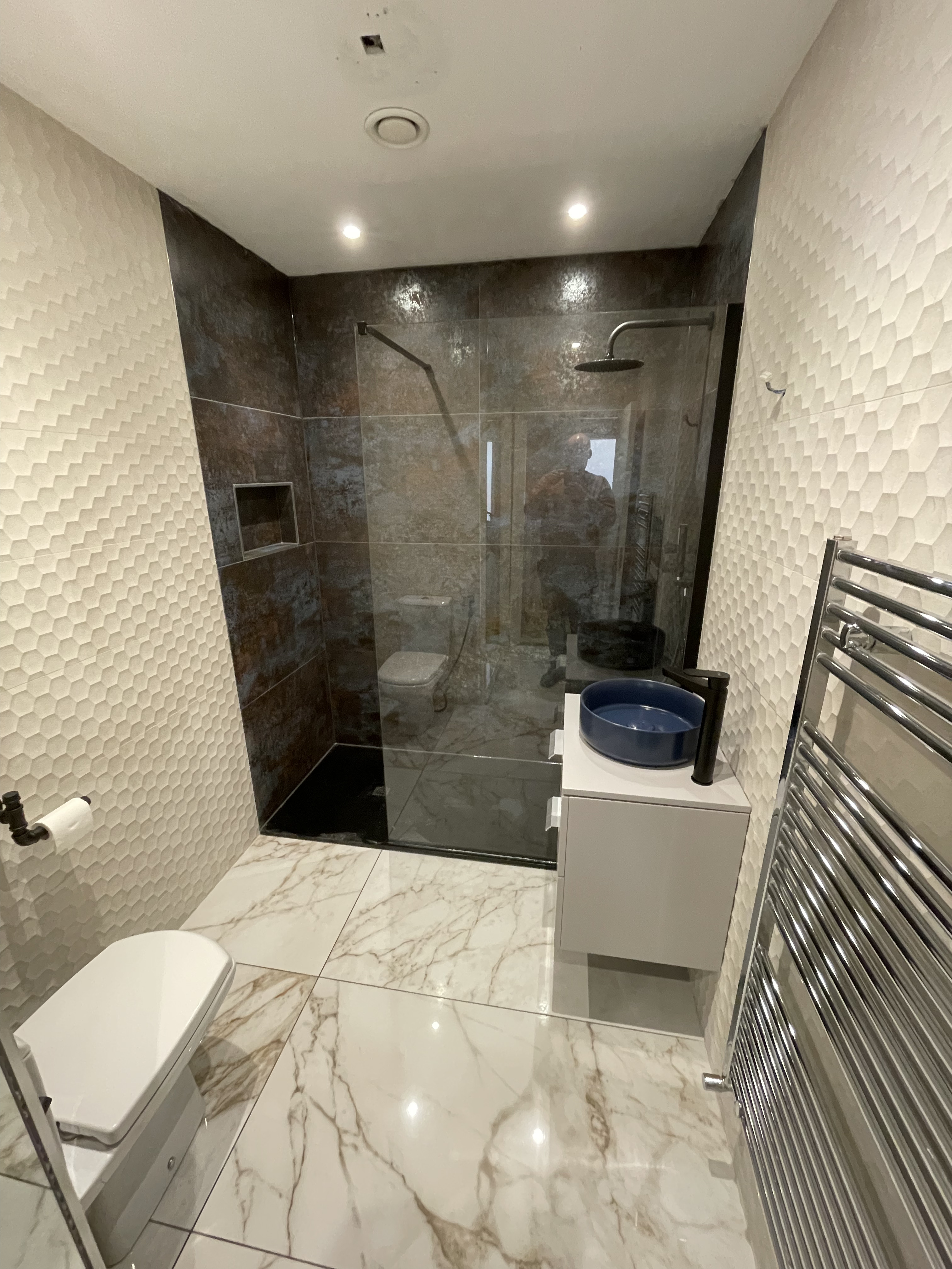 Custom bathroom with dark brown and blue hued tiled shower with a glass screen behind a white tiled bathroom with a circle sink and towel heater on the right and toilet on the front left.