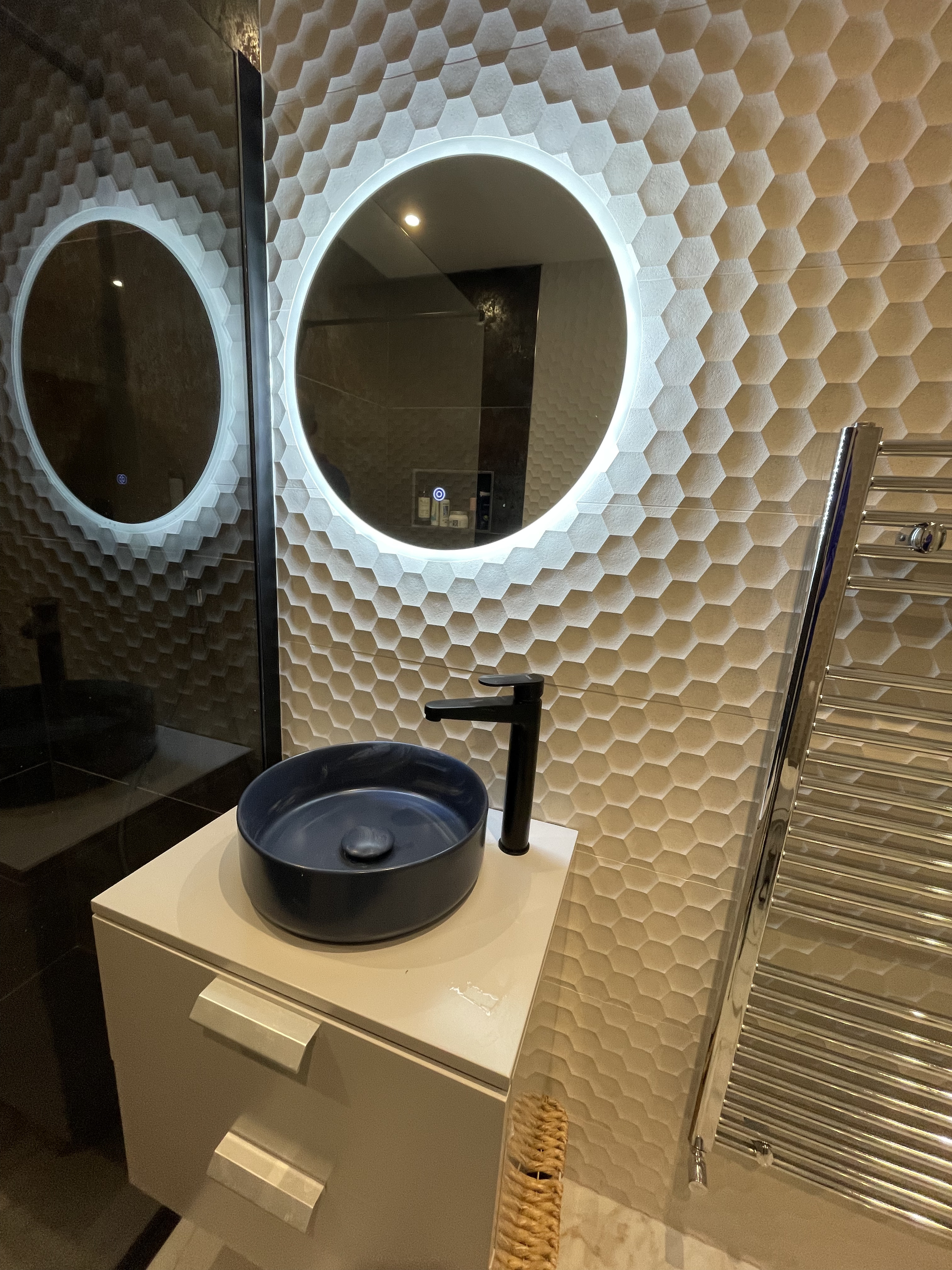White honeycomb backdrop behind a framless backlit circle mirror over a black circle sink with a stainless steel towel heater to the right