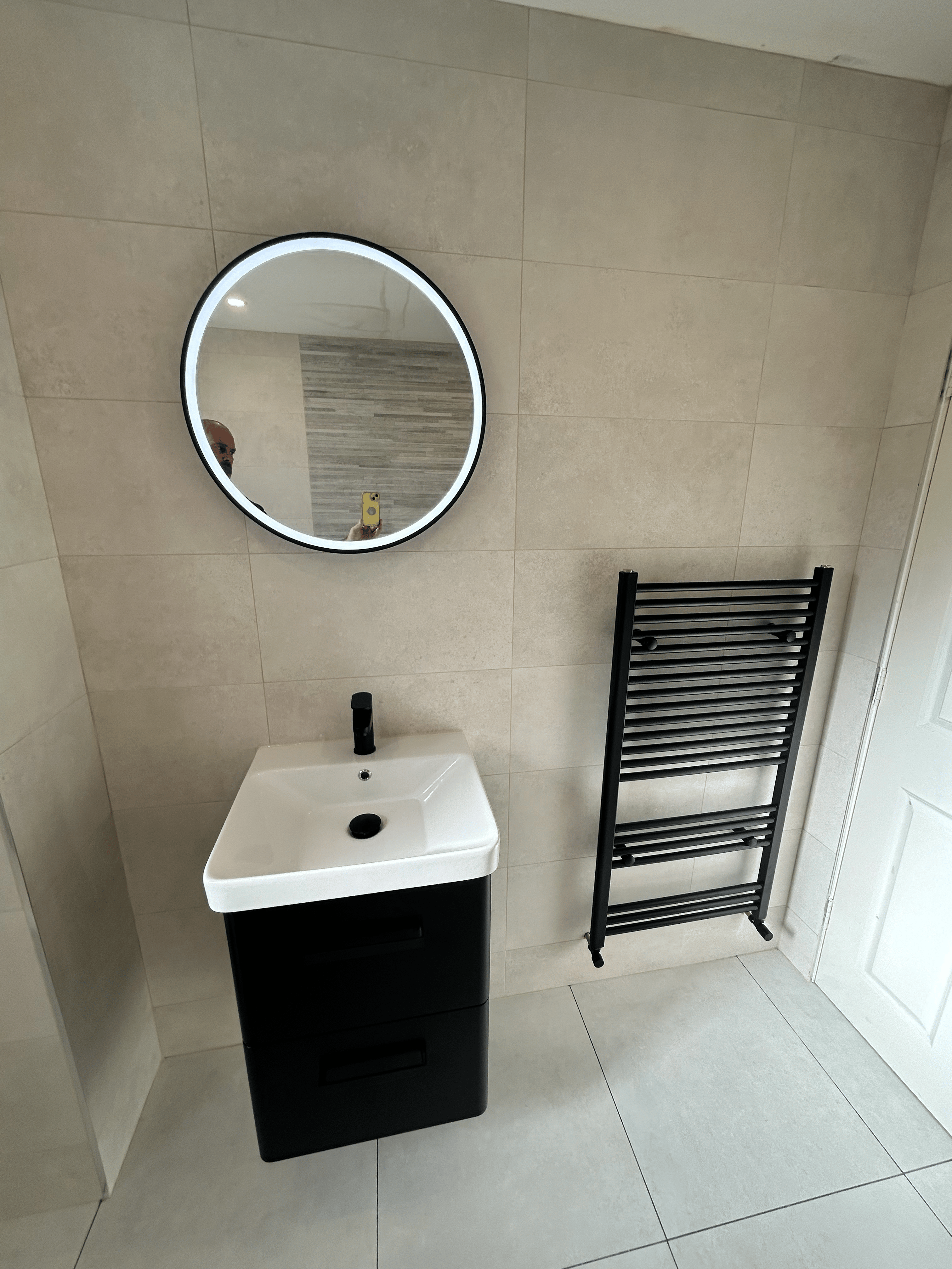 Light beige backwall of a bathrom with a white square sink under a lit circle mirror and black towel heater to the right.