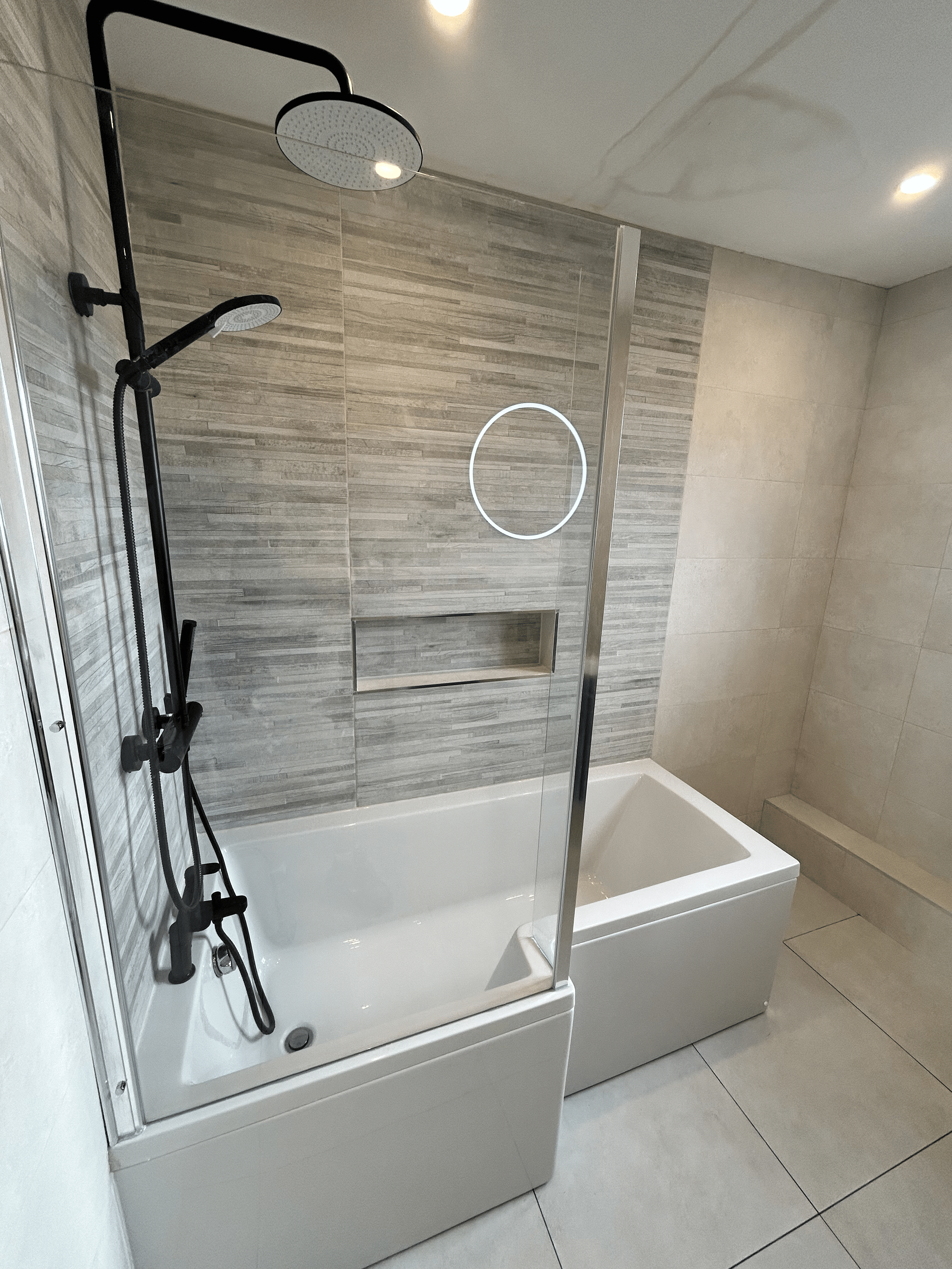 Grey tiled backwall with a cut out hole beside a white bathtub. Glass screen in the front with black shower head seen in the left